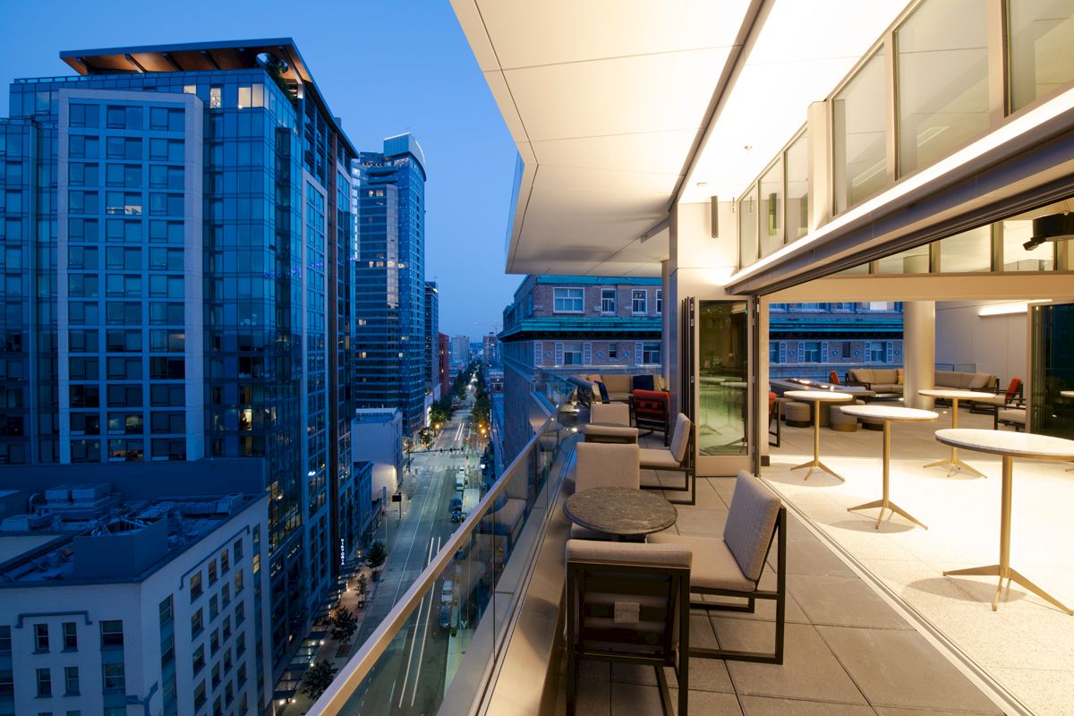 A modern cityscape at dusk with tall buildings and an elegantly furnished balcony overlooking the street.