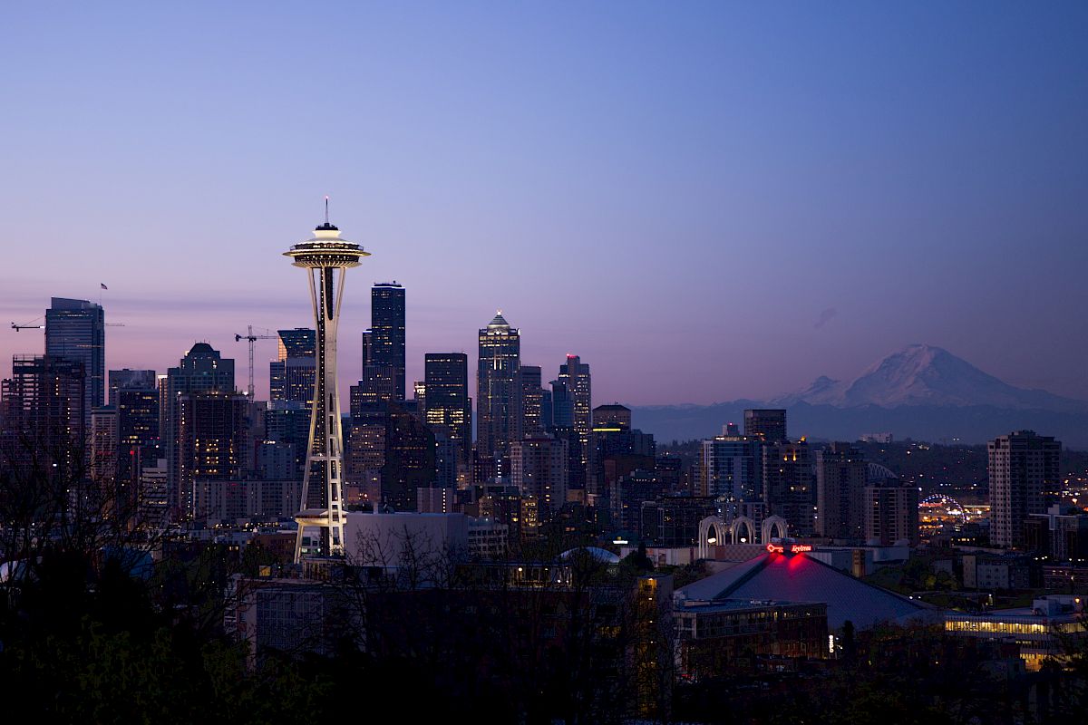 The image shows a city skyline with the Space Needle, skyscrapers, and Mount Rainier in the background during twilight.