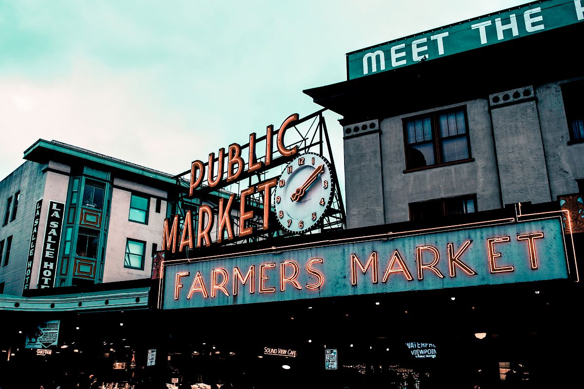 The image shows signs for a "Public Market" and "Farmers Market" with a clock, on a building under cloudy skies.