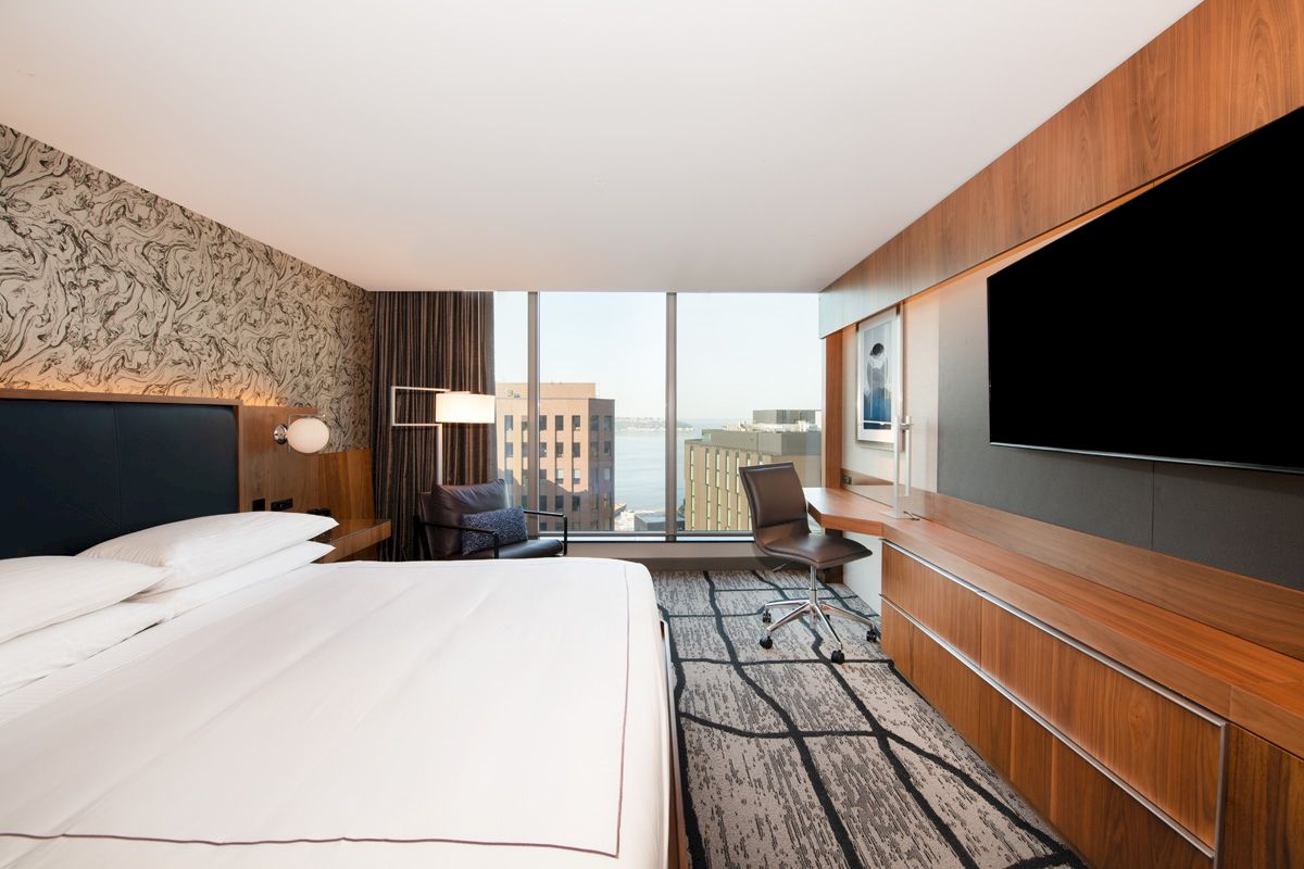 A modern hotel room with a large bed, wall-mounted TV, desk, and a window view of buildings outside.