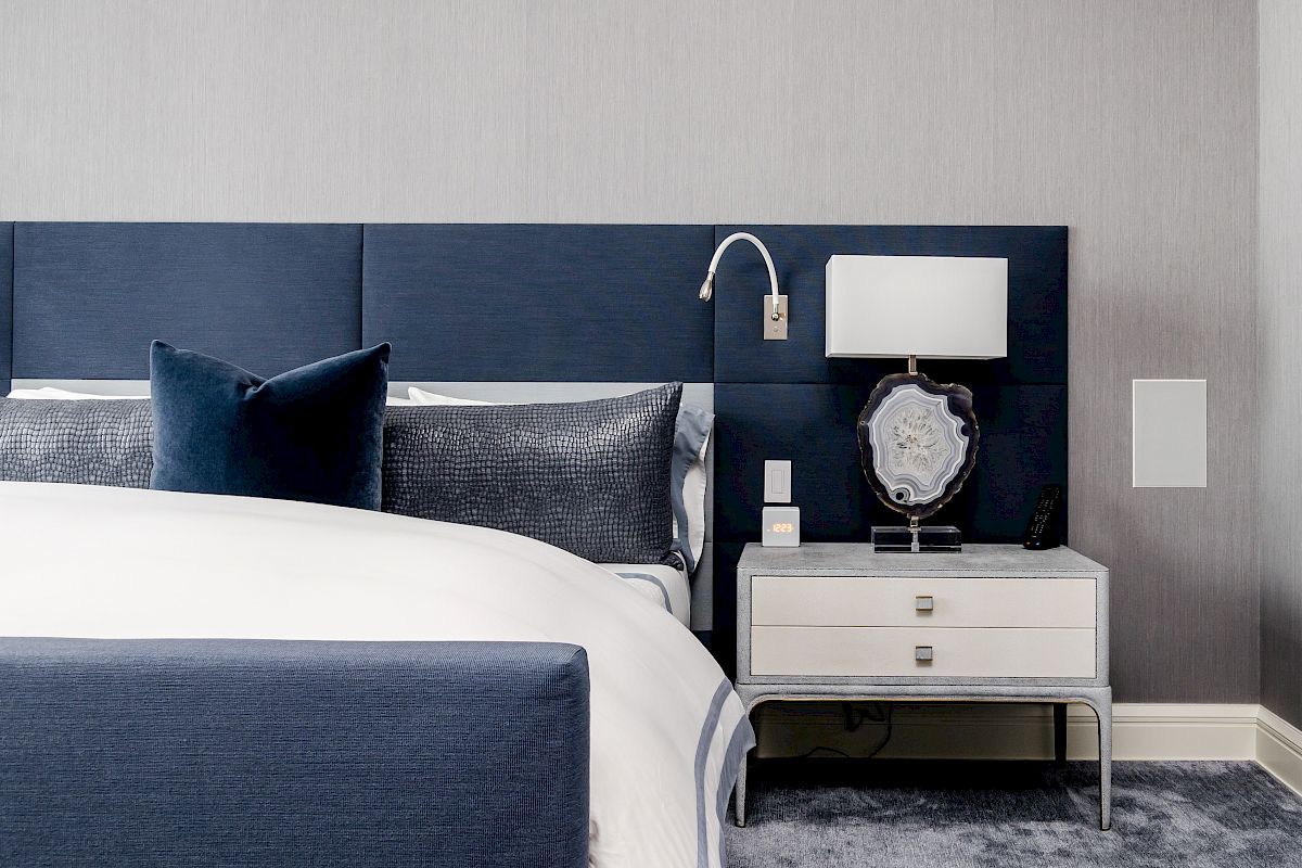 A modern bedroom with a blue headboard, white bedding, nightstand, lamp, and decor on a gray carpet and wall.