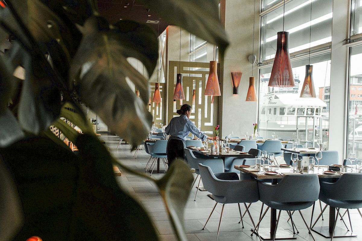 The image shows a restaurant interior with modern decor, a server setting tables, and warm lighting from pendant lamps by large windows.