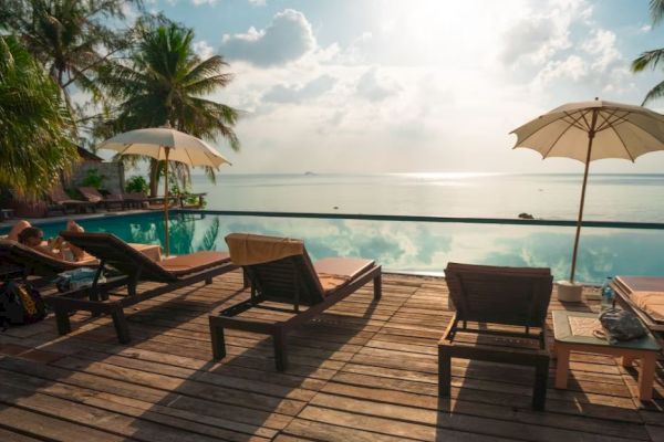 A serene poolside scene with lounge chairs, umbrellas, palm trees, and a view of a tranquil body of water under a bright sky.