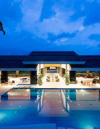 A luxurious building with a large, illuminated outdoor pool at dusk, surrounded by seating and greenery under a vibrant blue sky.