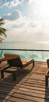 A serene poolside scene with lounge chairs, umbrellas, and palm trees, overlooking a calm body of water under a bright sky.