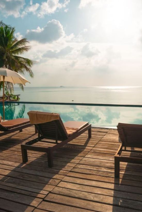 Poolside view with lounge chairs and umbrellas on a wooden deck, overlooking the ocean with palm trees and a bright sky above.