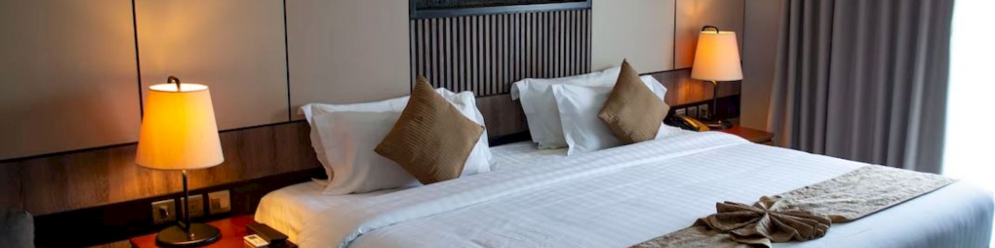 A neatly arranged hotel room features a large bed with brown and white bedding, bedside lamps, and a wall-mounted decorative element.