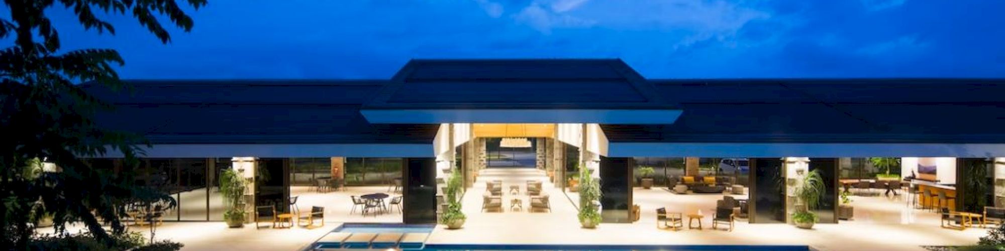 A luxurious building beside a lit swimming pool at dusk, under a blue sky with scattered clouds, surrounded by trees.