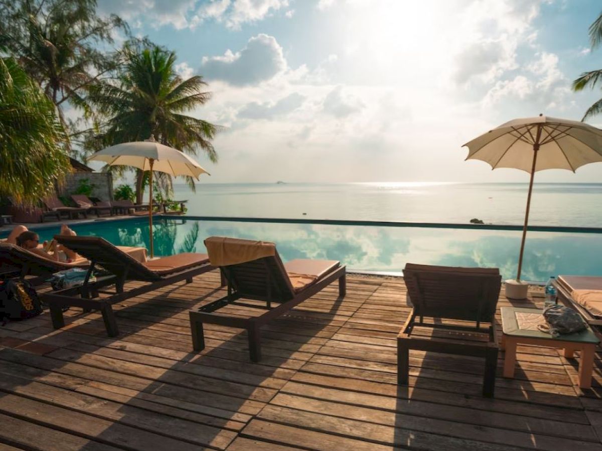 A serene poolside scene with lounge chairs, umbrellas, palm trees, and a view of the ocean under a bright sky.