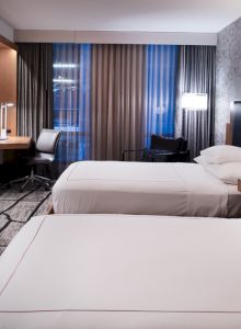 A modern hotel room with two beds, a TV, desk, and lamps, featuring patterned wallpaper and carpet.