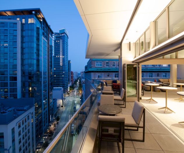 A modern balcony overlooks a city street with tall buildings, featuring outdoor seating and a view of the urban skyline at dusk.