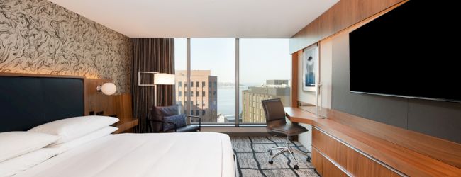 A modern hotel room with a large bed, wood paneling, a desk, chair, and a window with a city view is shown.