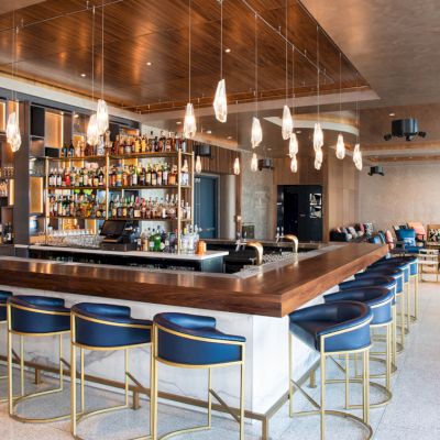 The image shows a modern bar with blue stools, hanging lights, and shelves stocked with bottles, adjacent to a cozy seating area.