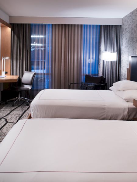 A modern hotel room with two double beds, a wall-mounted TV, a desk with a chair, contemporary lighting, and patterned wallpaper.