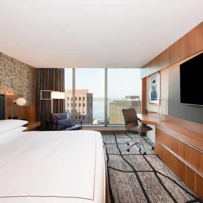 A modern hotel room with a large bed, desk, chair, TV, and a view of buildings through a wide window.