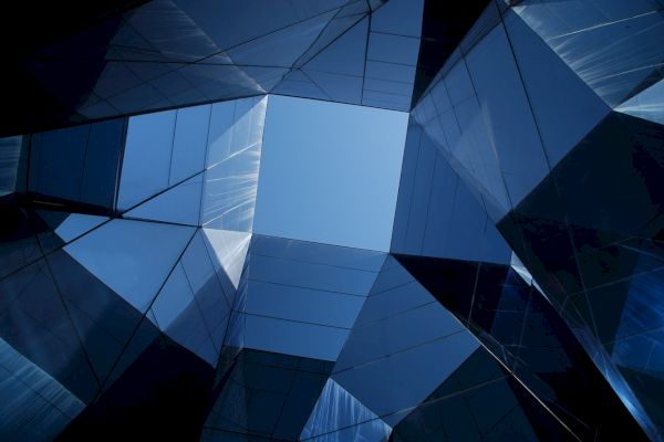 Architectural photo of tall mirrored buildings, reflecting the sky and each other, forming a geometric pattern around an open sky.