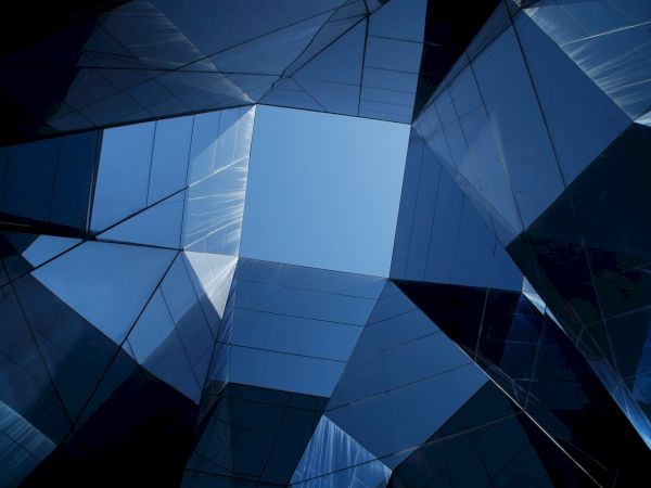 Architectural photo of tall mirrored buildings, reflecting the sky and each other, forming a geometric pattern around an open sky.