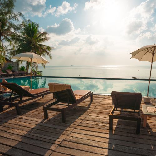 Lounge chairs and umbrellas by a pool, overlooking the ocean. Palm trees and a tranquil setting under a partly cloudy sky.