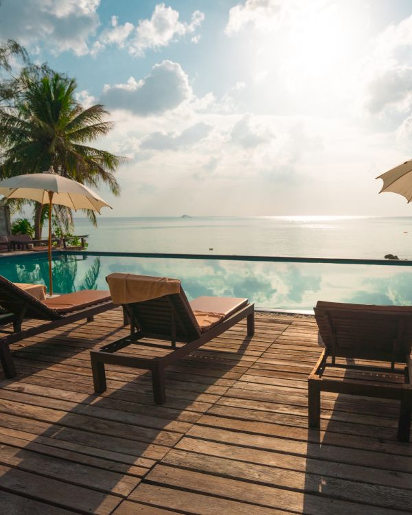 Lounge chairs and umbrellas by a pool, overlooking the ocean. Palm trees and a tranquil setting under a partly cloudy sky.