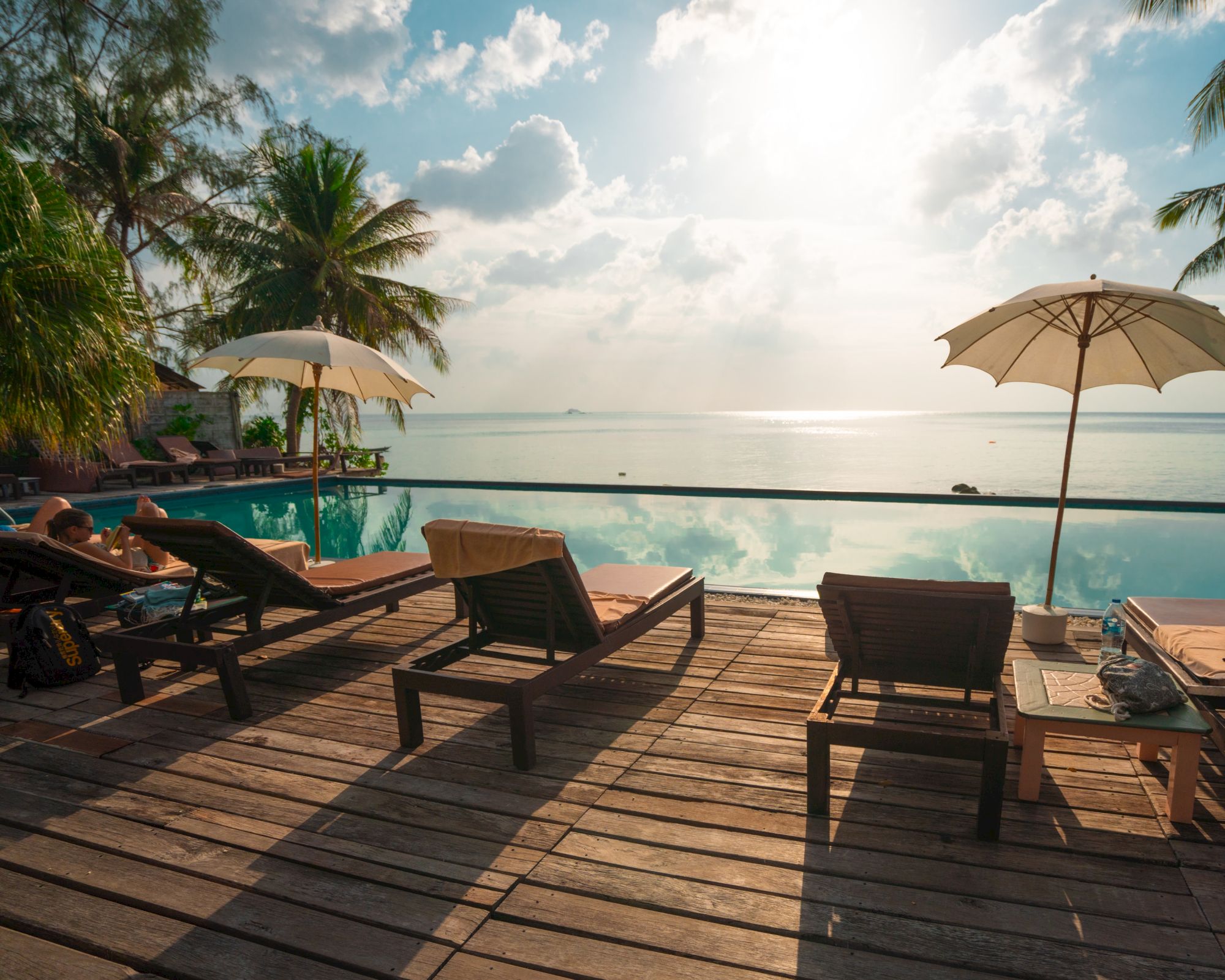 A serene poolside scene with loungers and umbrellas overlooks a calm ocean, framed by palm trees and a bright sky.