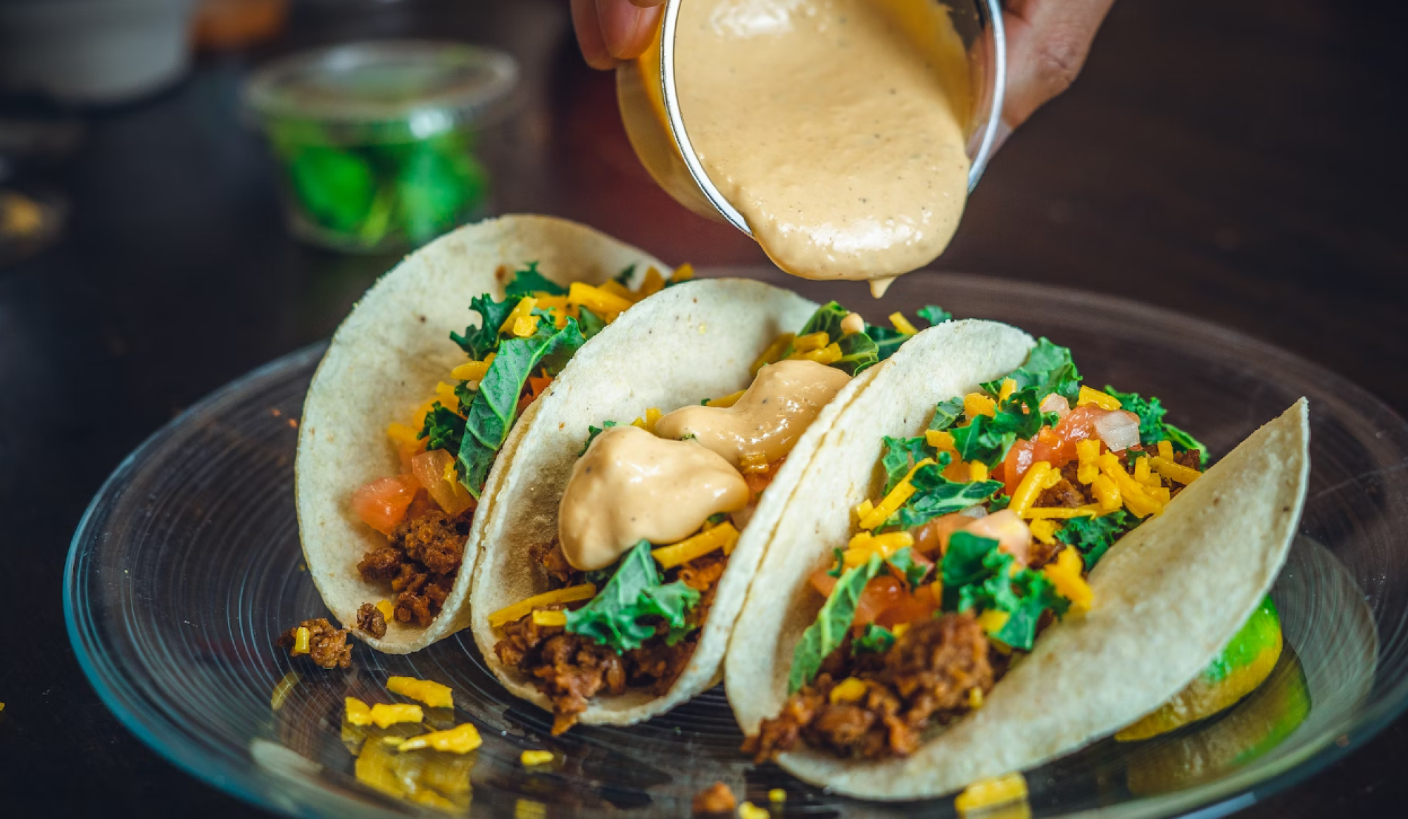 Three tacos on a plate with lettuce, cheese, and sauce being poured, garnished with small bowls of green garnish in the background.