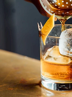 A hand pours a cocktail into a glass with ice and a lemon twist garnish on a wooden surface.