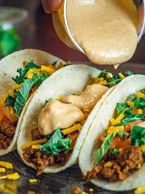 Three tacos with beef, lettuce, tomatoes, and cheese are topped with creamy sauce being poured from a container.