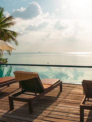 A serene poolside view with lounge chairs, umbrellas, palm trees, and an ocean backdrop. The scene is peaceful and sunlit.