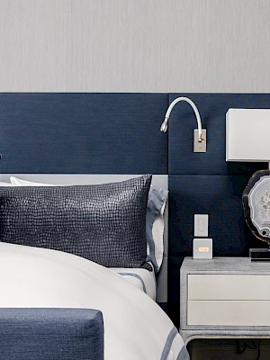 A neatly designed bedroom with a dark blue headboard, bedside table with a lamp, and pillows on the bed complete the scene.