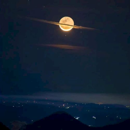 A bright moon appears ringed by clouds resembling Saturn, set against a dark sky over a distant illuminated landscape.