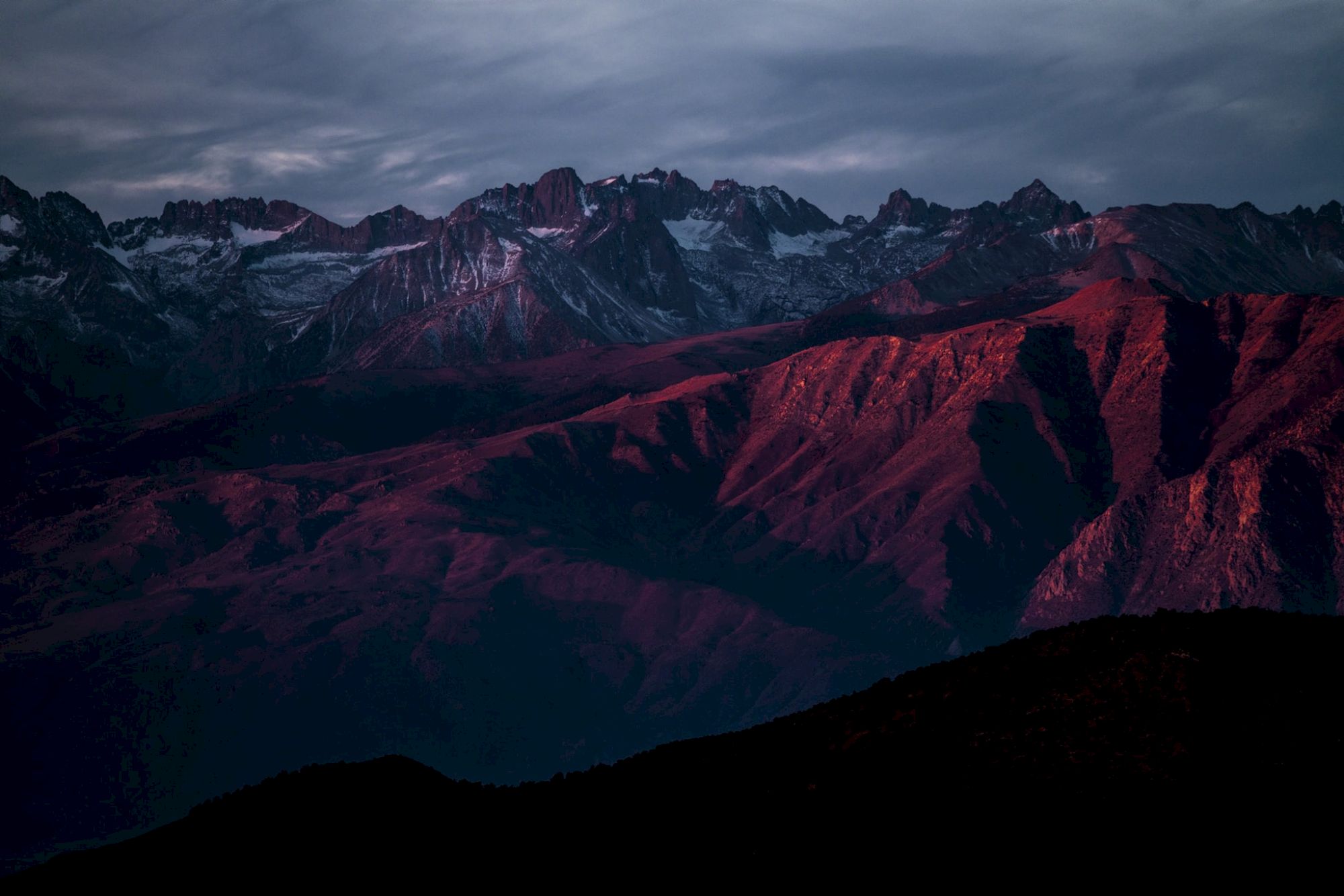 A mountain range with snow-capped peaks is bathed in red light under a cloudy sky, creating a dramatic landscape scene.