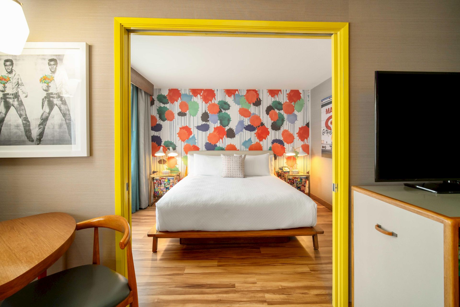 A modern bedroom with colorful polka dot wallpaper, a white bed, wooden floor, bright yellow door frame, and a TV to the right.
