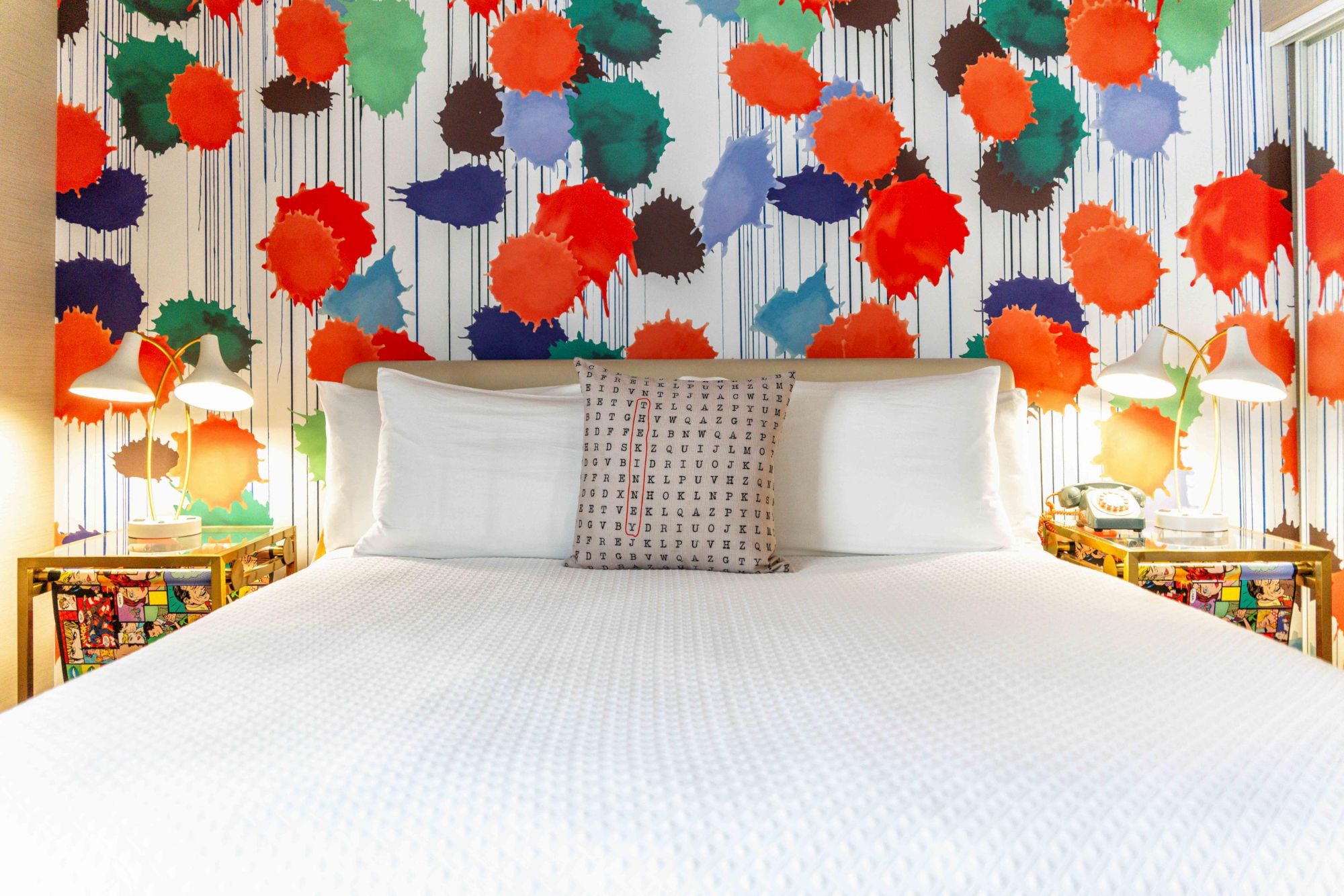 A bed with white bedding, colorful abstract wall art, bedside lamps, and a decorative pillow is shown in the image.
