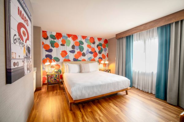 A brightly decorated bedroom features a bed, colorful accent wall, large window with teal curtains, and retro-style art.