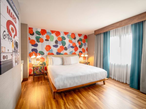 A brightly decorated bedroom features a bed, colorful accent wall, large window with teal curtains, and retro-style art.