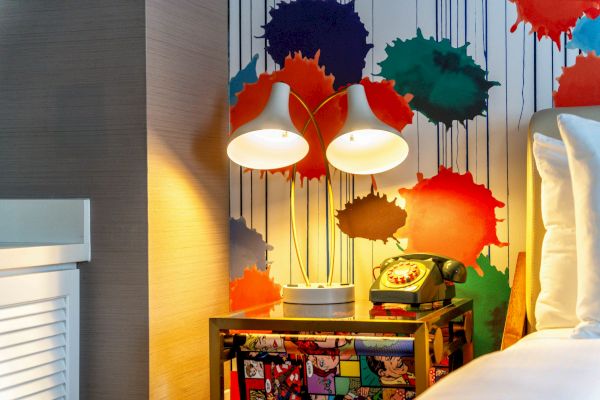 A colorful room corner with two lamps, a retro phone on a patterned table, and a vivid splatter wall art behind it.