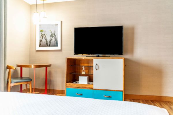 A modern hotel room with a TV, cabinet, small table, chair, and a framed picture on the wall is shown.