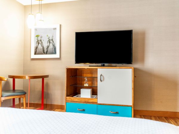 A modern hotel room with a TV, cabinet, small table, chair, and a framed picture on the wall is shown.