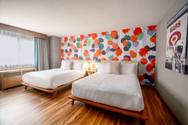 A hotel room with two beds, colorful floral wallpaper, a window with curtains, wooden floor, and a Mars-themed poster on the wall.