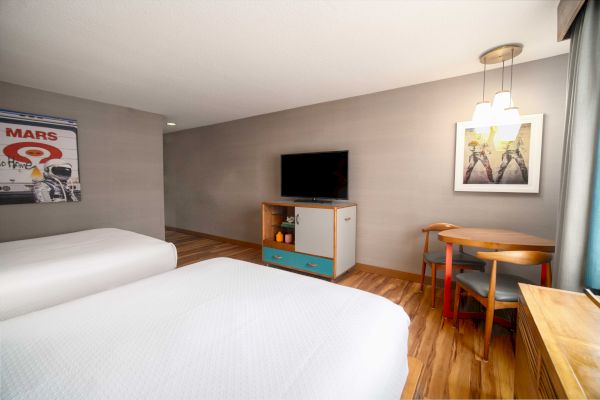 The image shows a modern hotel room with two beds, a TV, a small table with chairs, and wall art.