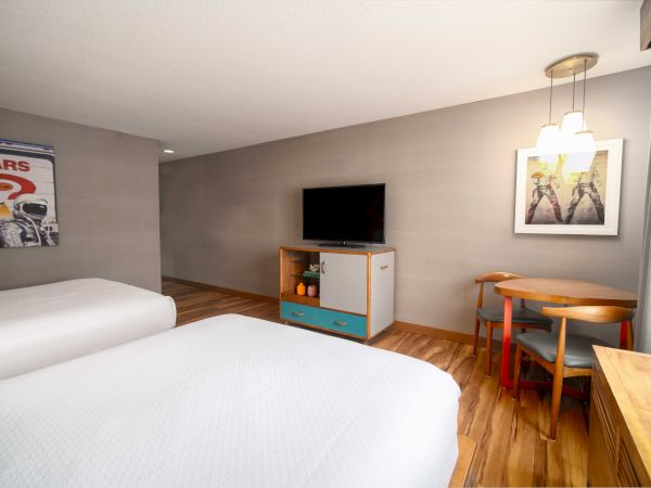 The image shows a modern hotel room with two beds, a TV, a small table with chairs, and wall art.