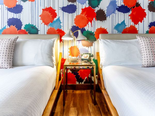A colorful bedroom with two beds, vibrant paint splatter wallpaper, and a nightstand with a retro lamp and telephone between them.