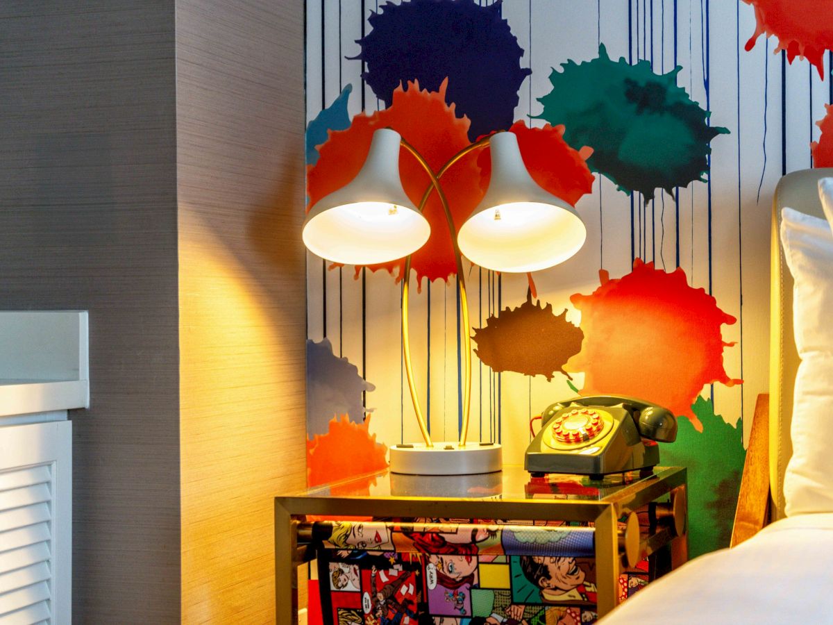 A colorful room with a nightstand featuring pop art, two modern lamps, a vintage phone, and a vibrant splatter wall design.