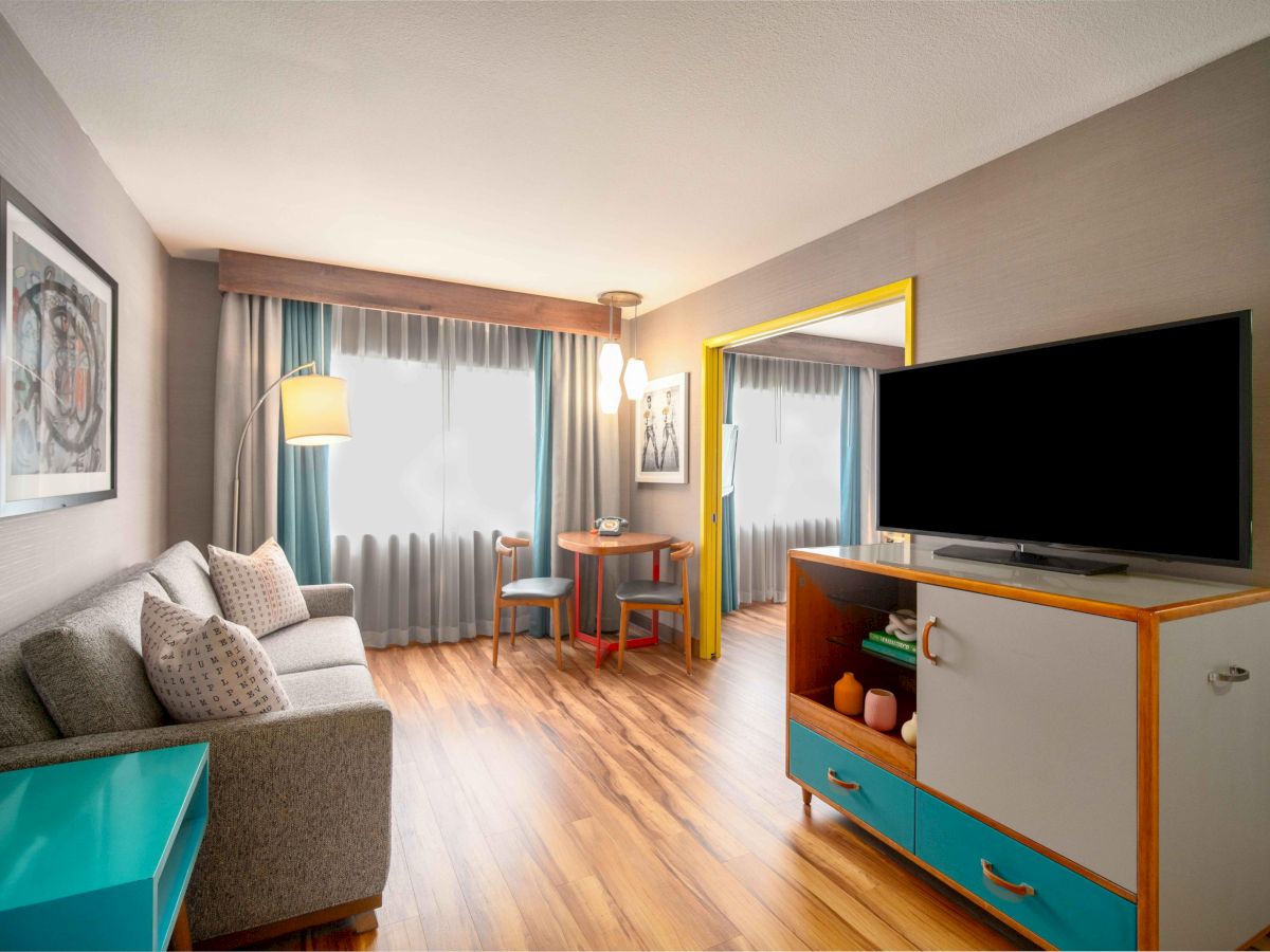 The image shows a modern living room with a sofa, TV on a colorful stand, a small table with chairs, a lamp, and large window curtains.