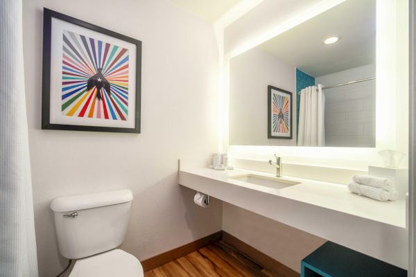 A bathroom with modern decor: a toilet, illuminated mirror, sink, towel rack, and colorful artwork on the wall complete the space.