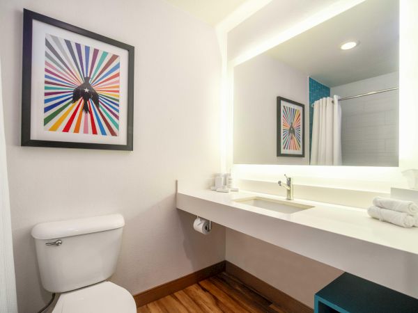 A bathroom with modern decor: a toilet, illuminated mirror, sink, towel rack, and colorful artwork on the wall complete the space.
