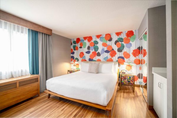 A modern bedroom with a colorful, abstract wallpaper, a white bed, wooden flooring, and natural light from a window.