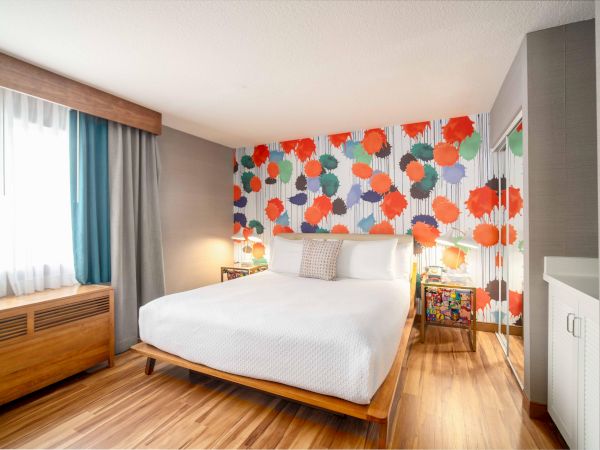 A modern bedroom with a colorful, abstract wallpaper, a white bed, wooden flooring, and natural light from a window.