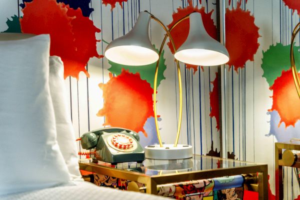 A bedside table with a vintage rotary phone, two modern lamps, and colorful splatter artwork on the wall behind it.