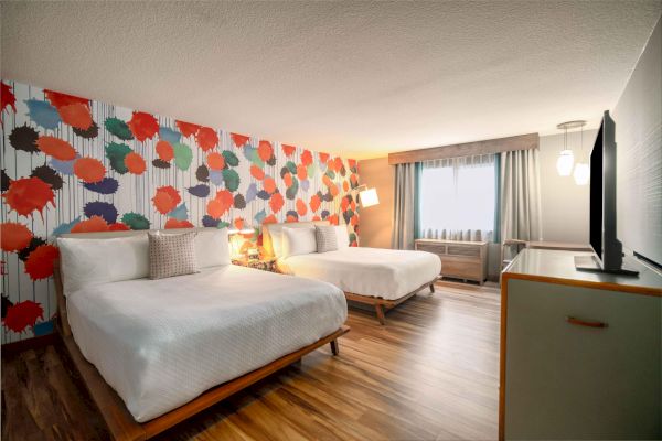 The image shows a hotel room with two beds, colorful wall art, wooden floor, a flat-screen TV, and a window with curtains.